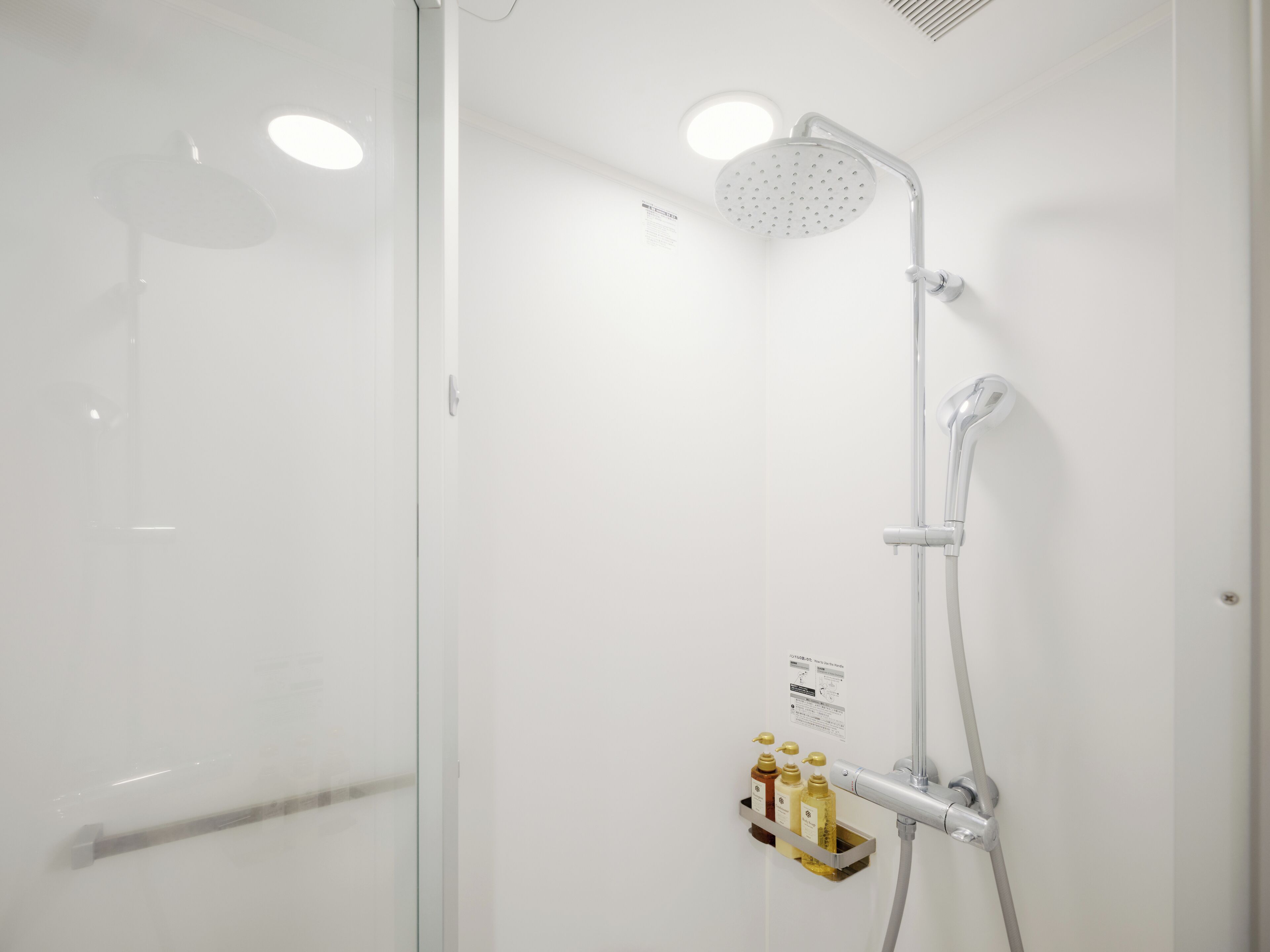 A modern shower with a handheld showerhead and a wall-mounted faucet. There is a control panel with instructions and a soap dispenser mounted on the wall.