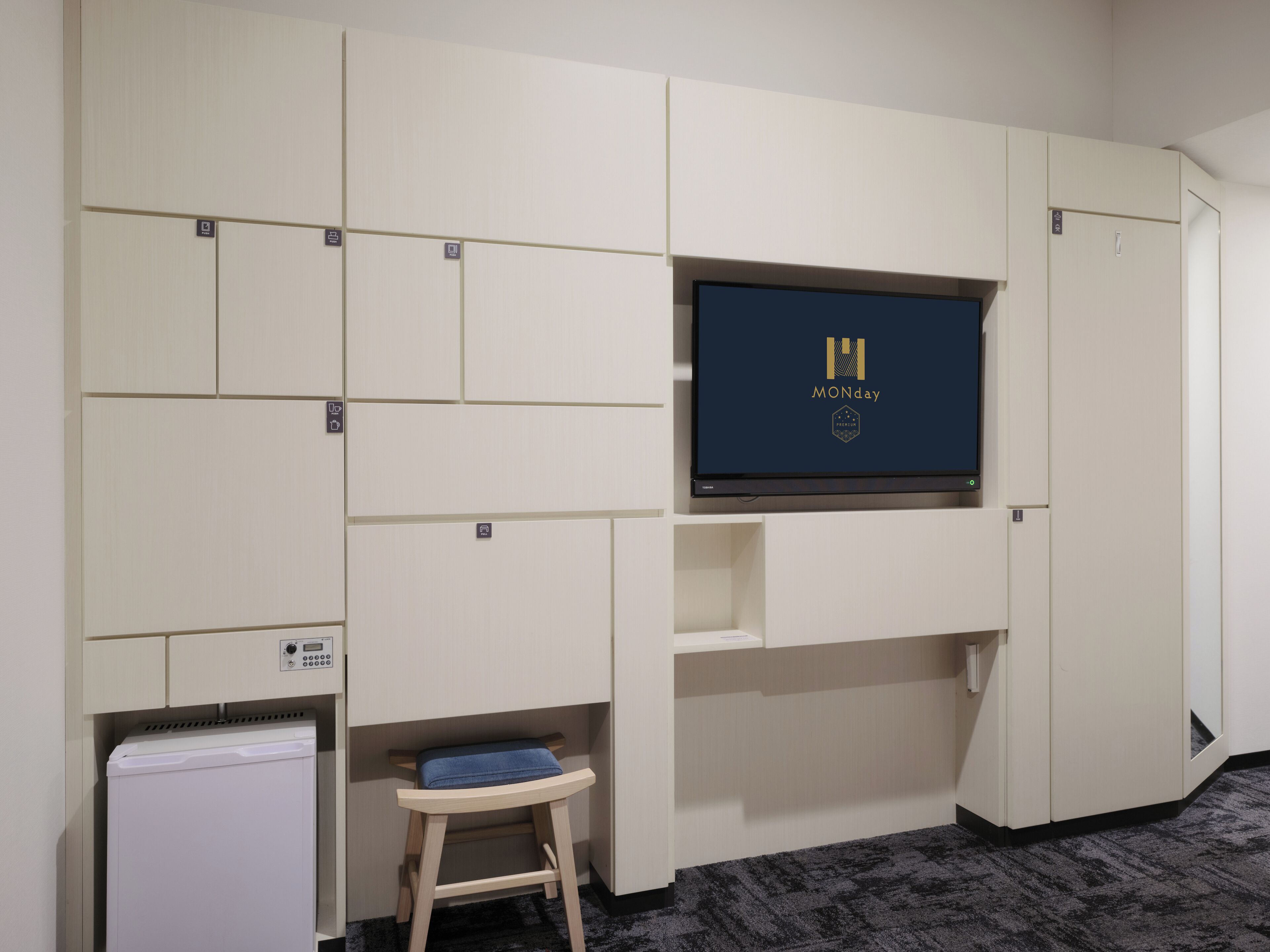 A hotel room with a flat-screen TV mounted on the wall, a small wooden stool, and a built-in mini-fridge.