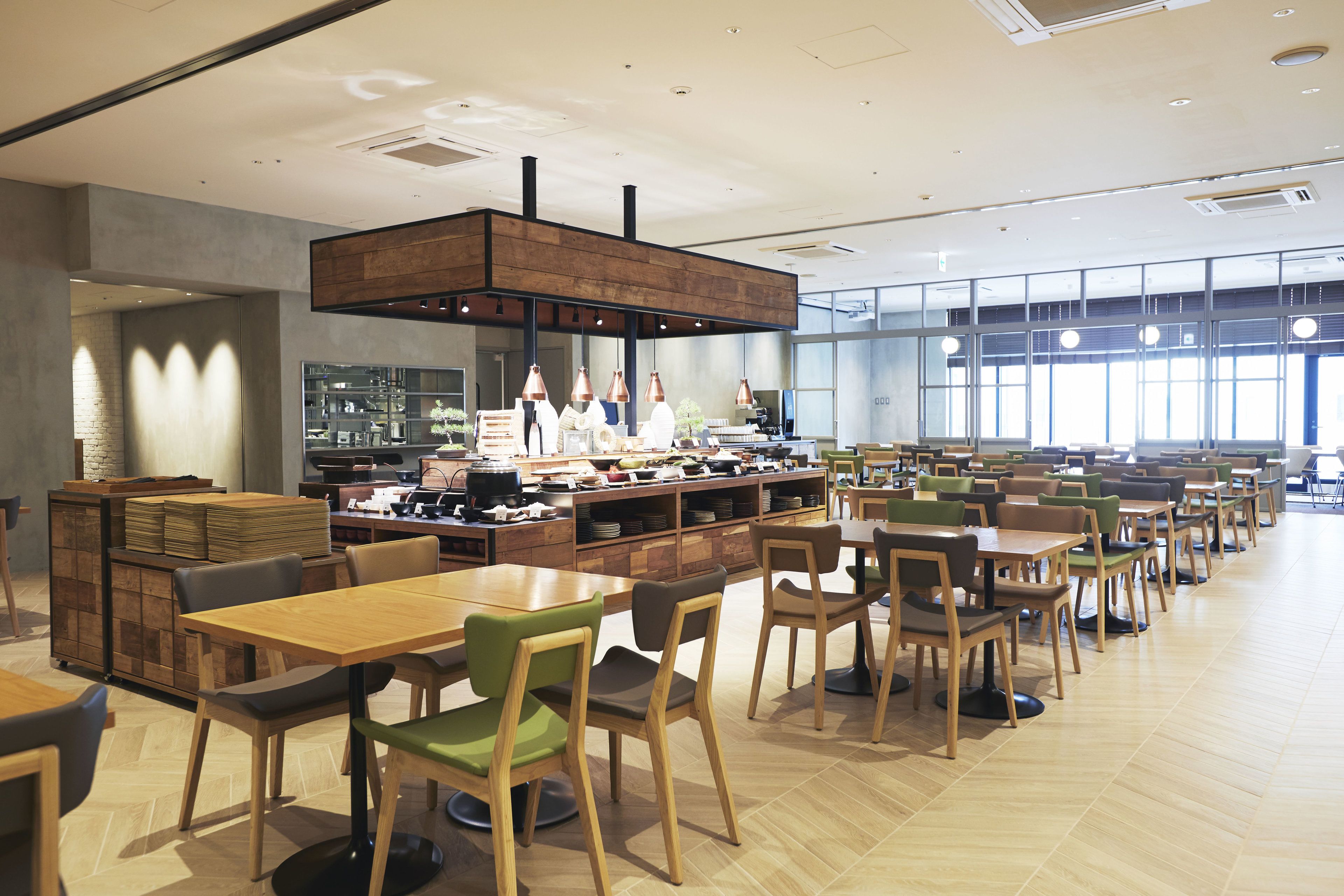 A spacious dining area with multiple tables and chairs, a central kitchen area with chefs, and large windows providing natural light.
