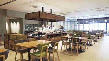 A spacious dining area with multiple tables and chairs, a central kitchen area with chefs, and large windows providing natural light.