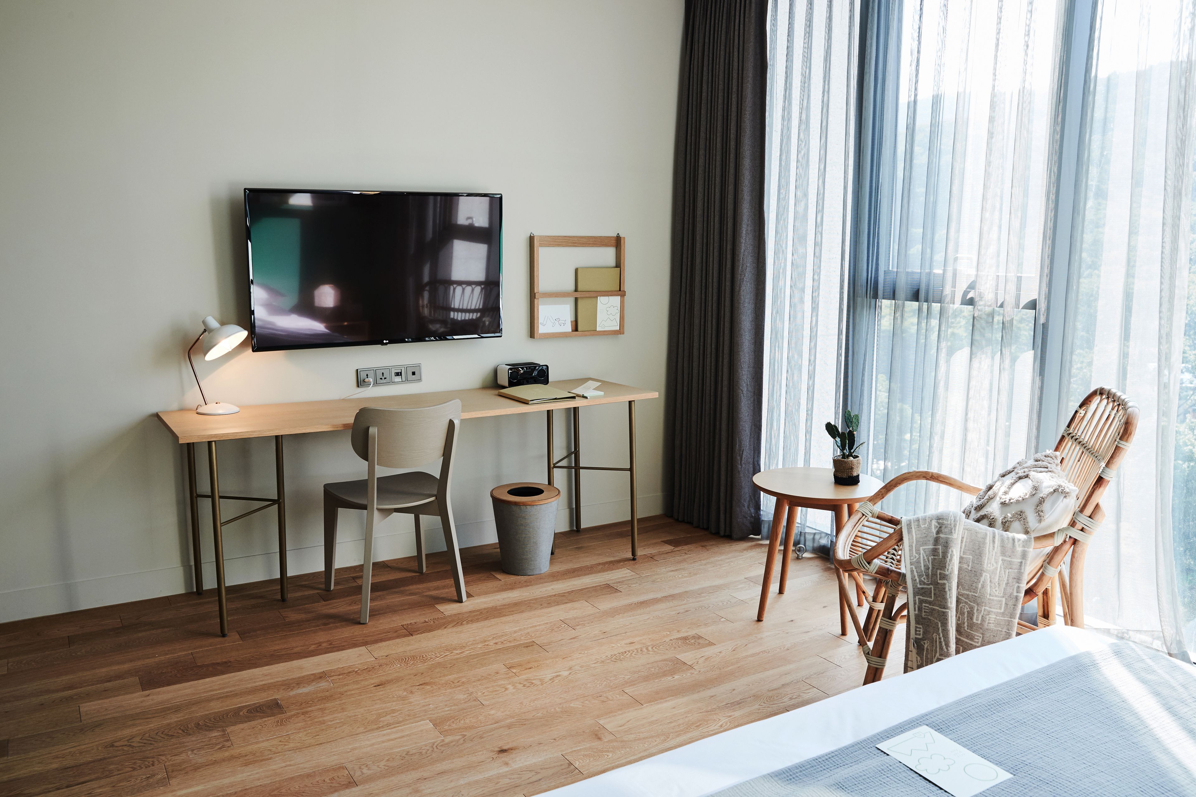 Una habitación de hotel moderna con un escritorio de madera, un televisor, una silla, una mesita con una planta y un ventanal grande con cortinas.