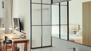 Una habitación de hotel moderna con un espejo grande, un escritorio con lámpara, una silla de madera, un lavamanos y un ventanal con persianas verticales.