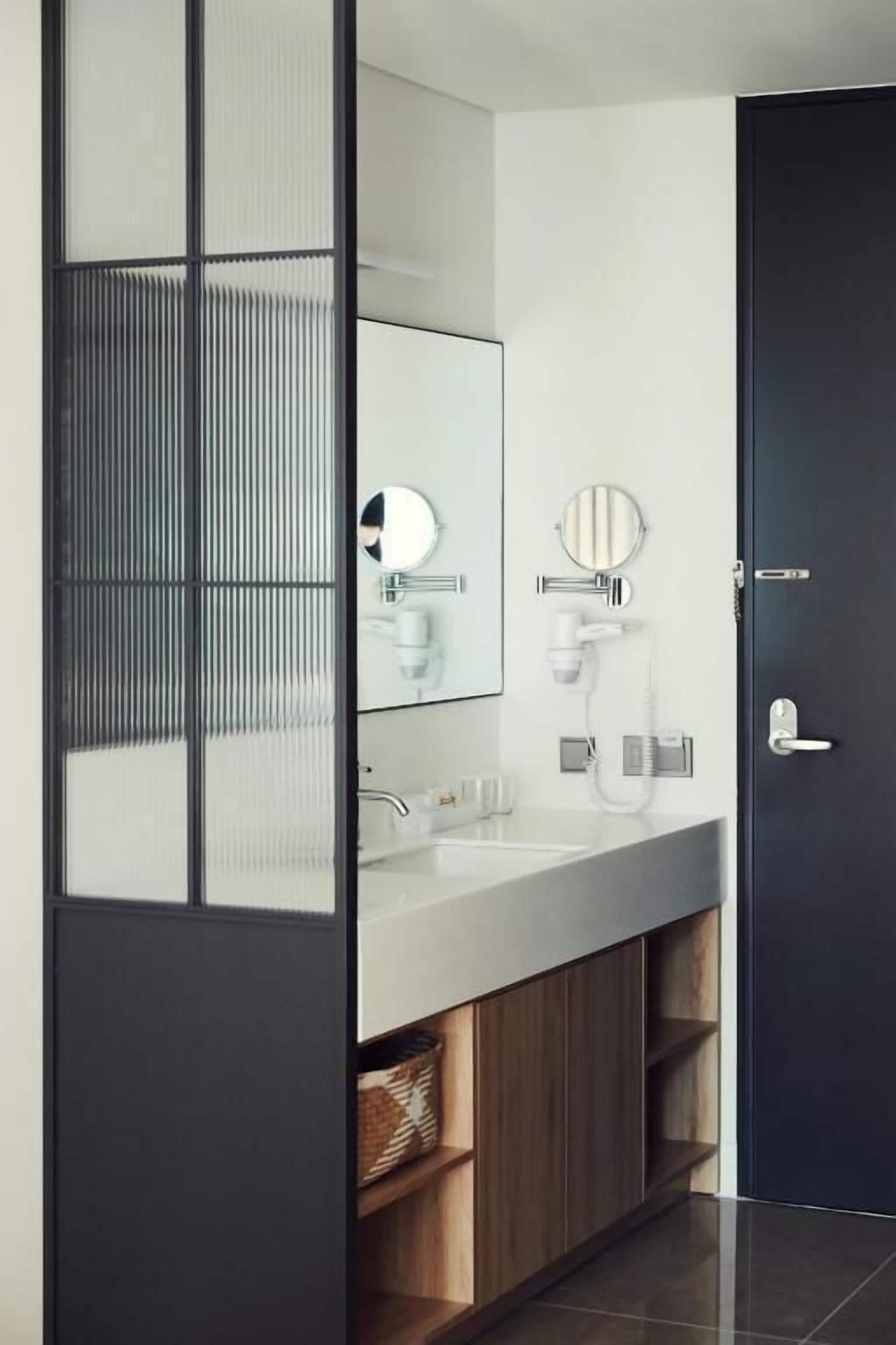 Un baño moderno con un espejo grande, un lavabo y un secador de pelo. Hay una puerta oscura y estantes de madera debajo del lavabo.