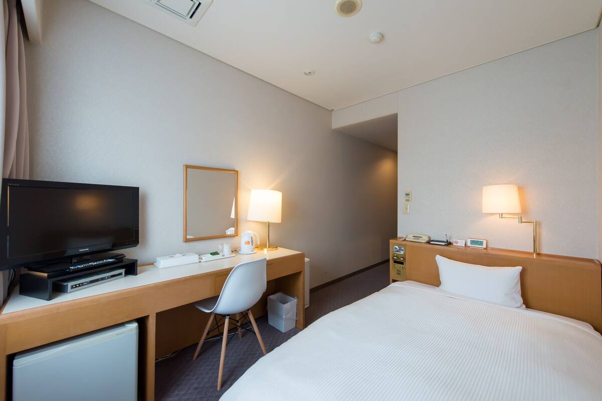 Habitación de hotel con cama, escritorio con televisor, silla, lámpara y mesita de noche con teléfono y reloj.