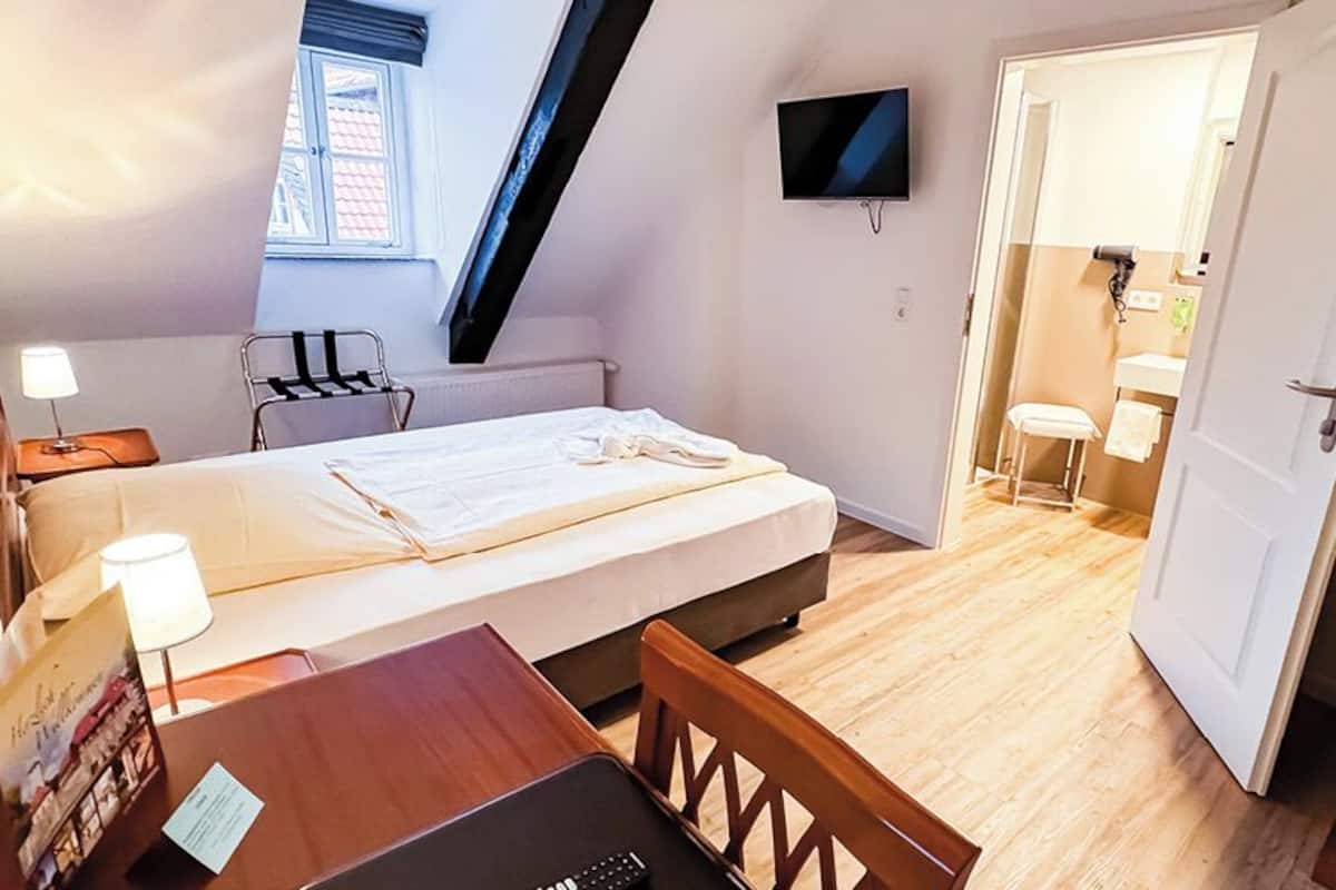 Een slaapkamer met een bed, een bureau, een stoel, een televisie en een zichtbare badkamer door een geopende deur.