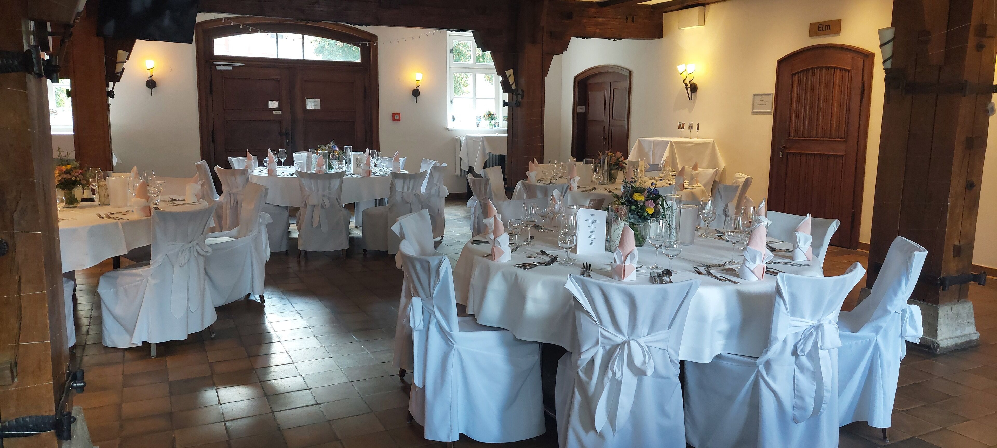 Ein Festsaal mit runden Tischen, gedeckt für eine festliche Veranstaltung, mit weißen Tischdecken, Stühlen mit Schleifen und floralen Tischdekorationen.