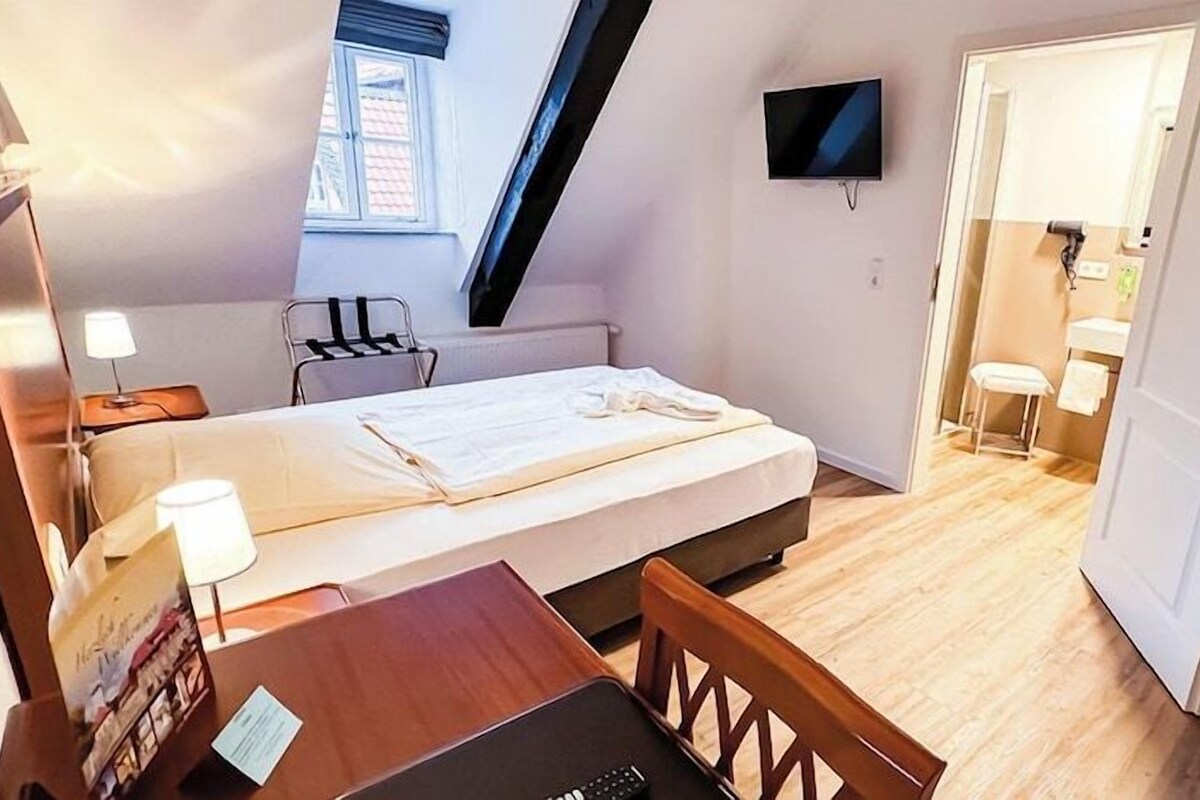 A bedroom with a bed, a desk with a laptop, a chair, a TV mounted on the wall, and a door leading to a bathroom.