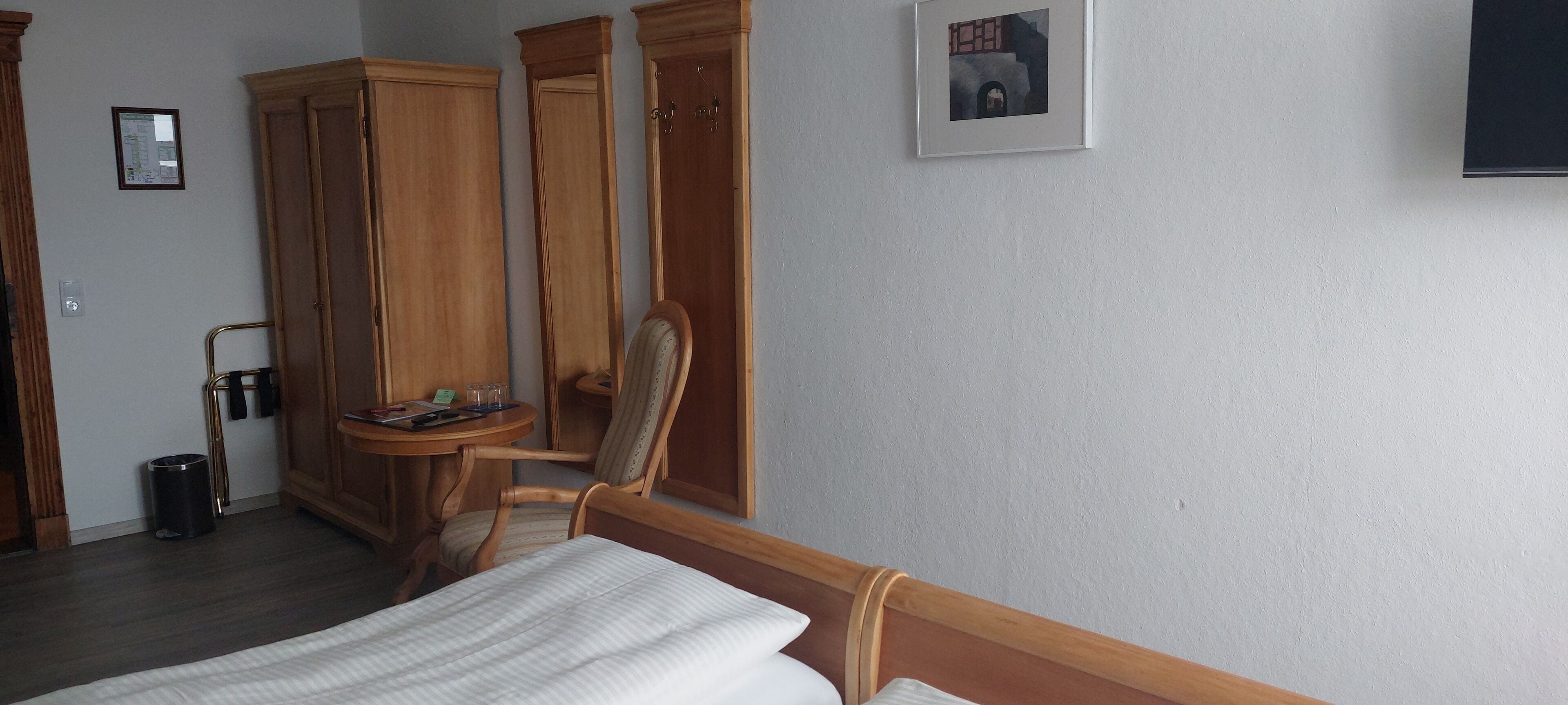 A hotel room with a bed, wooden wardrobe, a small table with a chair, a trash bin, and a framed picture on the wall.