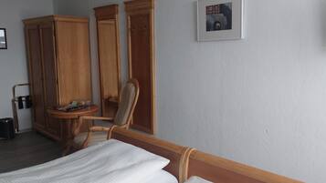 A hotel room with a bed, wooden wardrobe, a small table with a chair, a trash bin, and a framed picture on the wall.