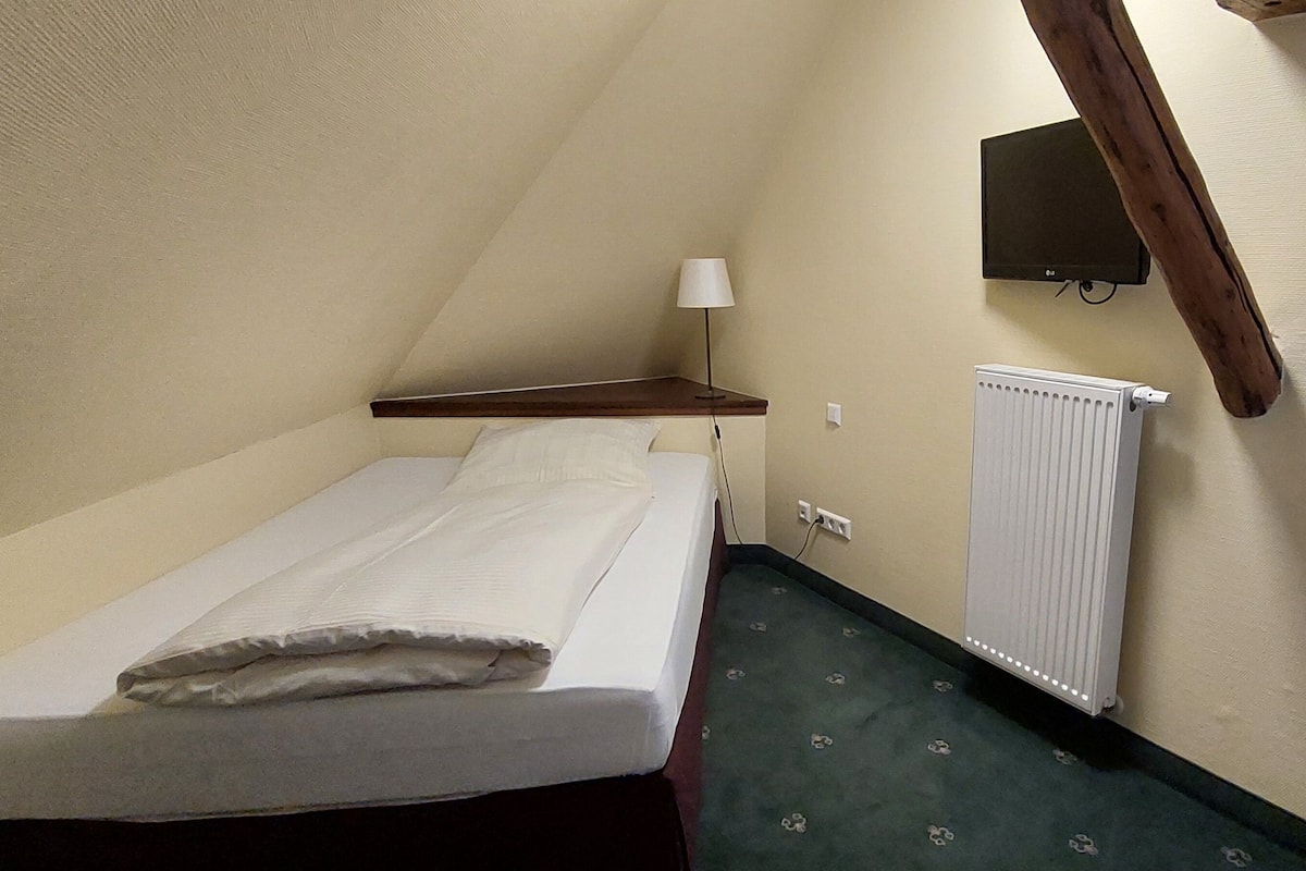 Een kleine eenpersoonskamer met een houten hoofdeinde, een nachtkastje met een lamp en een radiator aan de muur.