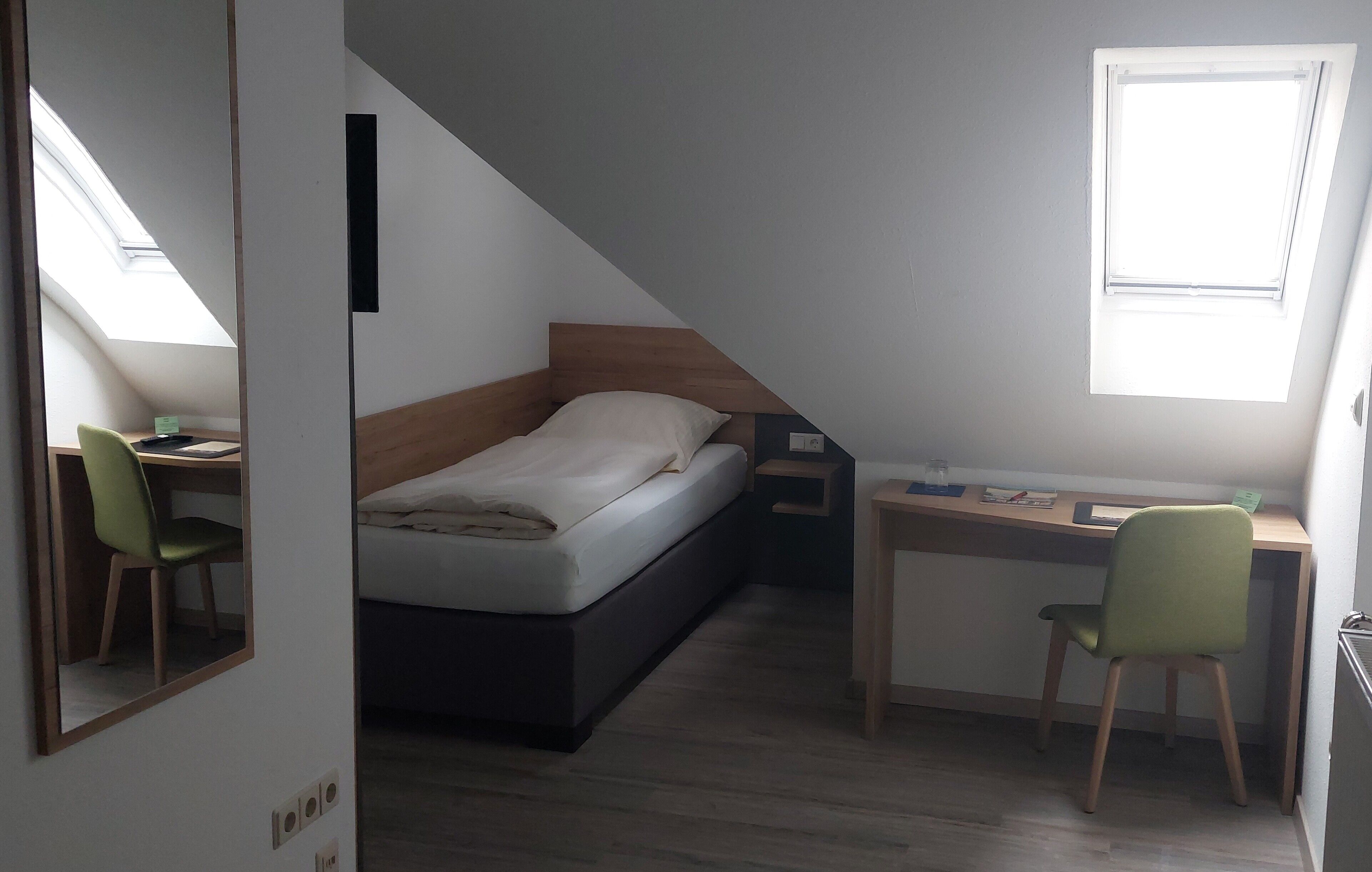 A compact room with a bed, a desk, and a chair. There is a skylight above the bed and a window on the desk side.