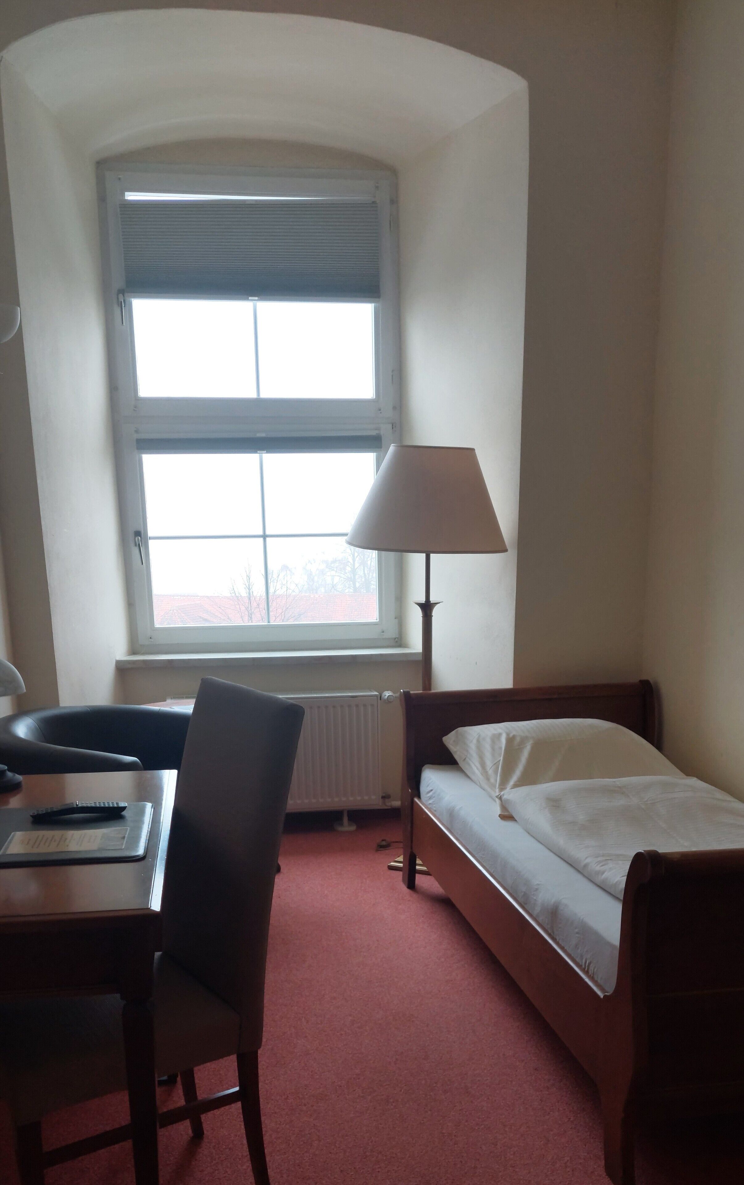 Una habitación pequeña y bien iluminada con una cama individual, un escritorio con silla, una lámpara y un ventanal con persianas.
