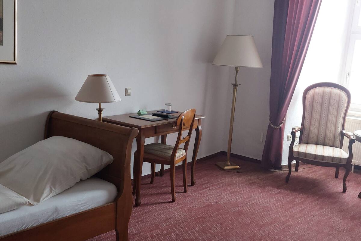 Een hotelkamer met een enkel bed, een bureau met stoel, een staande lamp en een raam met gordijnen.