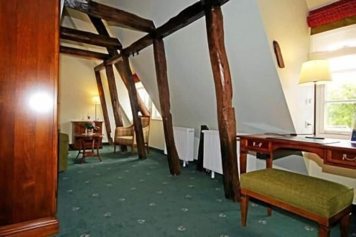 Een kamer met zichtbare houten balken, een bureau, een stoel en een raam met uitzicht op groen.