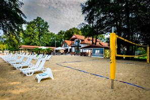 A beach with white lounge chairs lined up, a volleyball net, and a building with a red roof in the background.