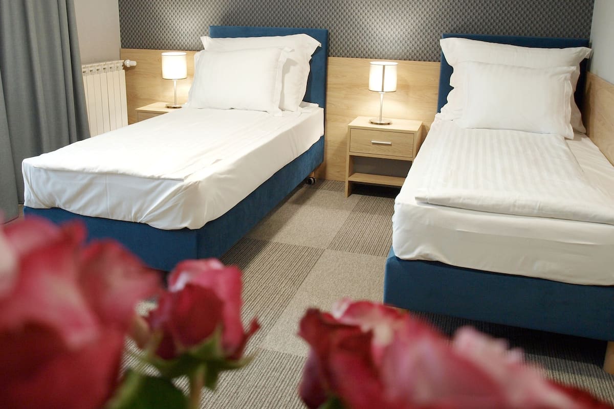 Hotelkamer met twee bedden, elk met een blauwe hoofdbord, witte beddengoed en bedlampjes. Er is een radiator aan de muur en een vloerbedekking.
