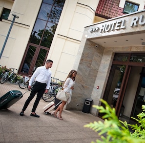 A hotel entrance with a sign, a man and a woman with luggage, and bicycles parked outside.