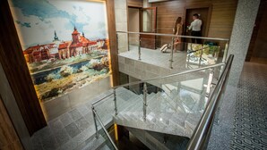 A multi-level interior with a glass railing, a large painting of a historical building, and a wooden door. - Hotel Rudnik (Grudziadz)