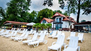 A beach with rows of white lounge chairs and a building with a red roof in the background. - Hotel Rudnik (Grudziadz)