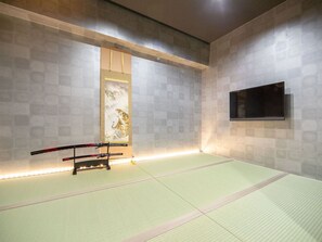 A tatami mat room with a mounted TV, a wooden shelf with a framed artwork, and a pair of swords on a stand.