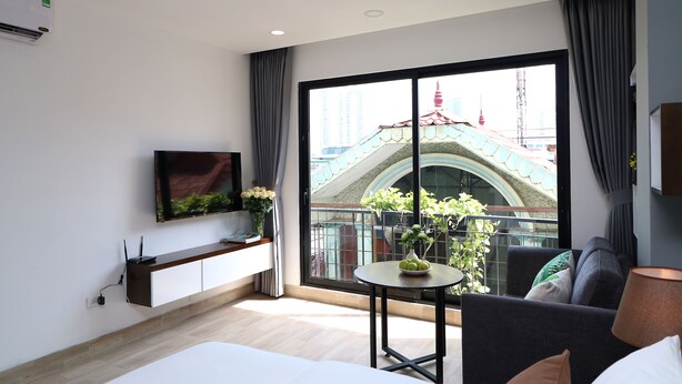 Een moderne hotelkamer met een groot raam, een flatscreen televisie aan de muur, een klein rond tafeltje met een bloevas, een bank en een lamp.