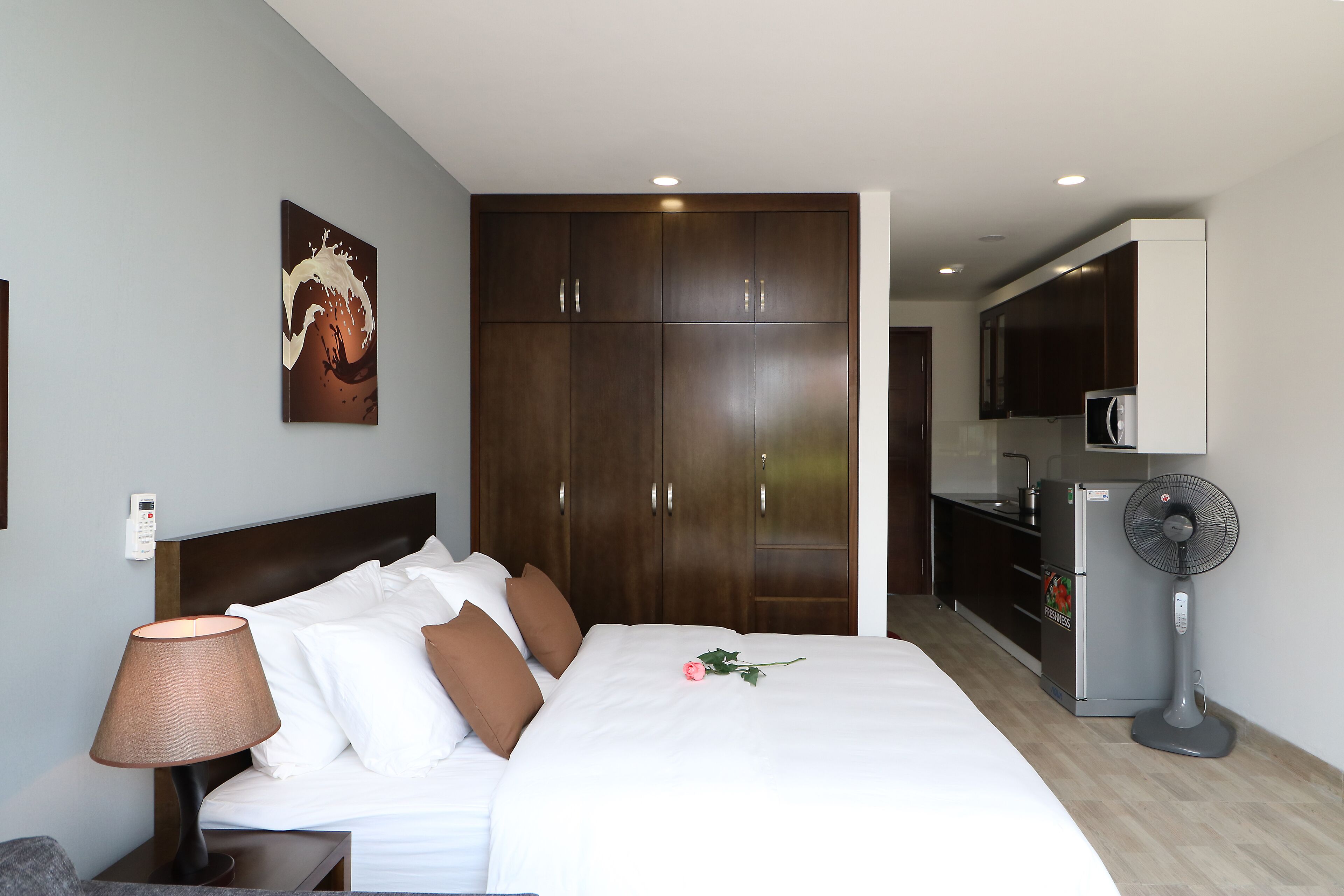 Habitación de hotel con cama, mesita de noche, lámpara, ventilador, microondas y kitchenette.