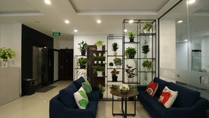 Een moderne lobby met twee blauwe banken, een glazen scheiding met plantenbakken, en een receptiebalie.