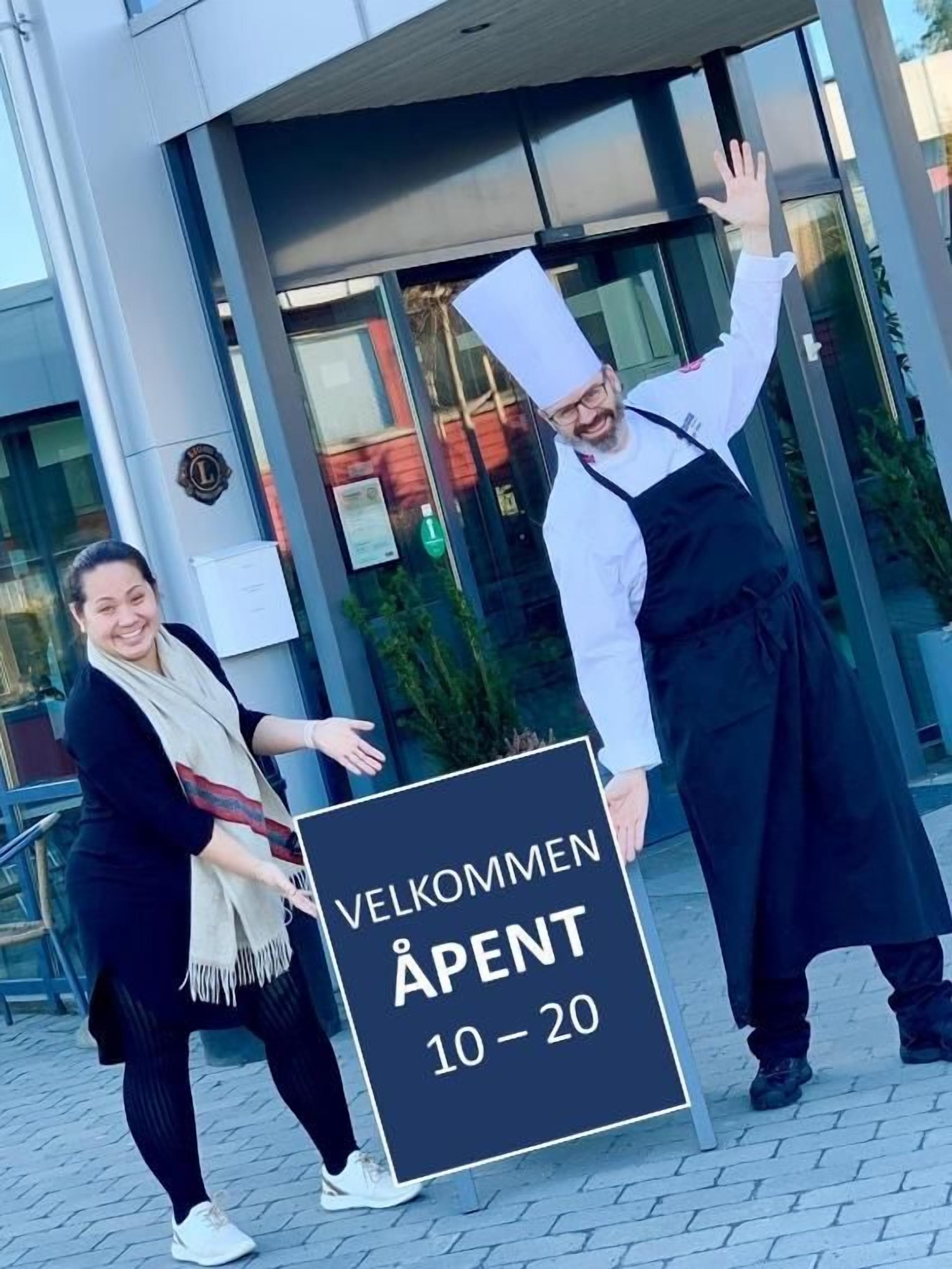 Two individuals holding a sign with 'VELKOMMEN ÅPENT 10-20' indicating a welcome event for rooms 10-20.