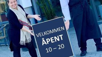 Two individuals holding a sign with 'VELKOMMEN ÅPENT 10-20' indicating a welcome event for rooms 10-20.
