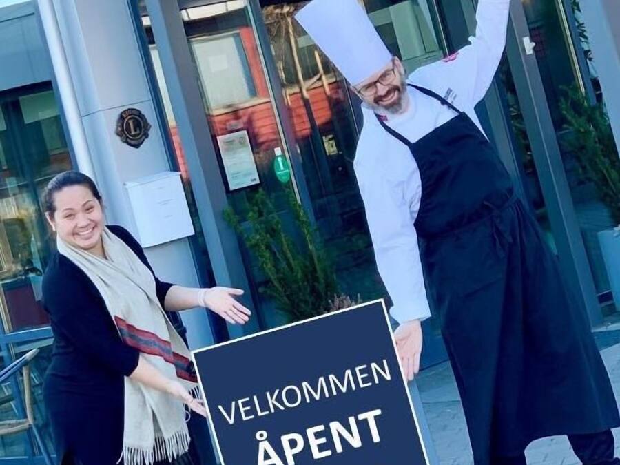 Two individuals holding a sign with 'VELKOMMEN ÅPENT 10-20' indicating a welcome event for rooms 10-20.