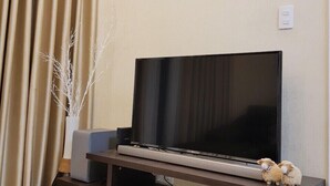 A living room with a flat-screen TV on a wooden stand, beige curtains, and a white vase with branches. - Florita SGN Green Living (Ho Chi Minh City)