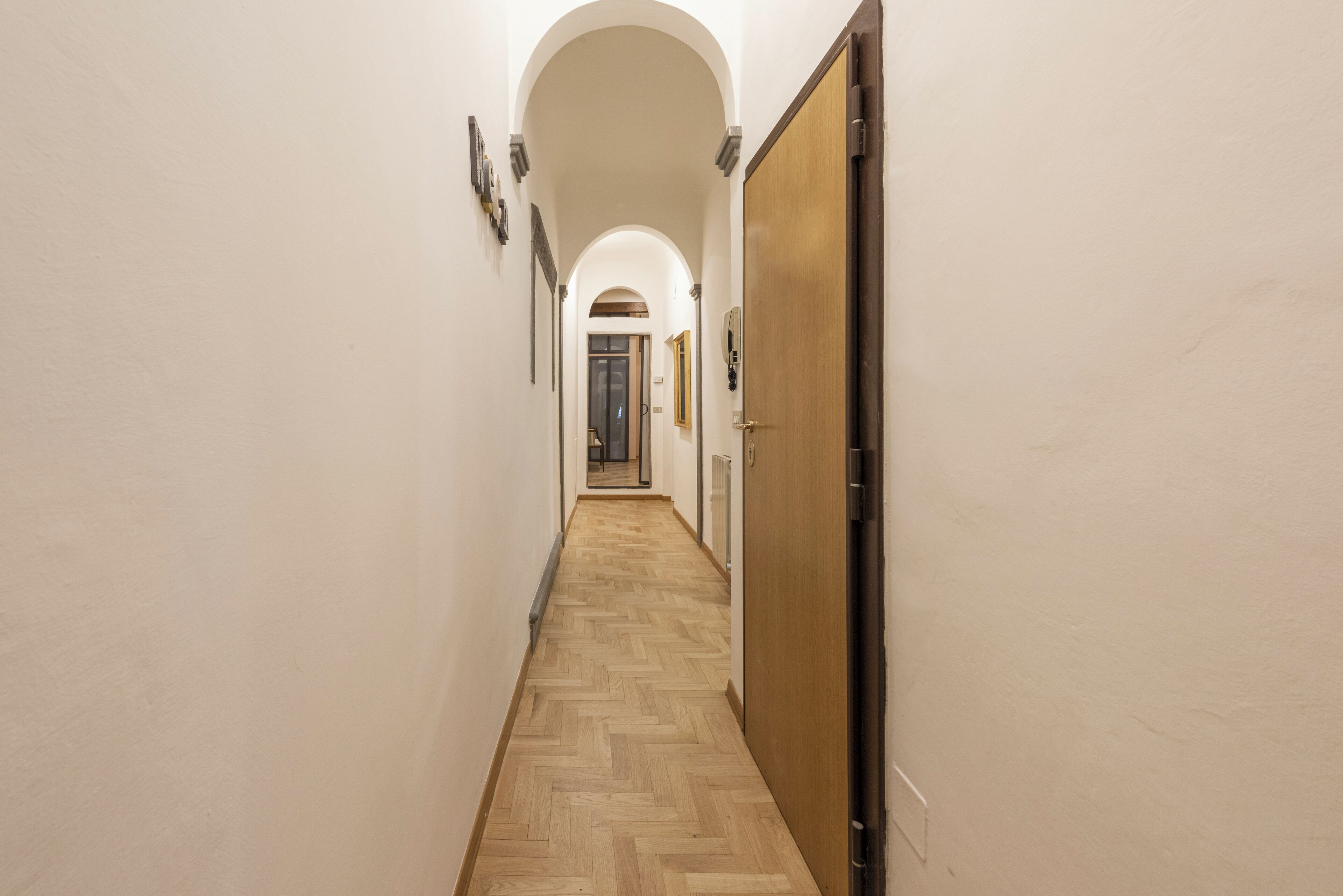 A hallway with a wooden herringbone floor, white walls, and a wooden door on the right.