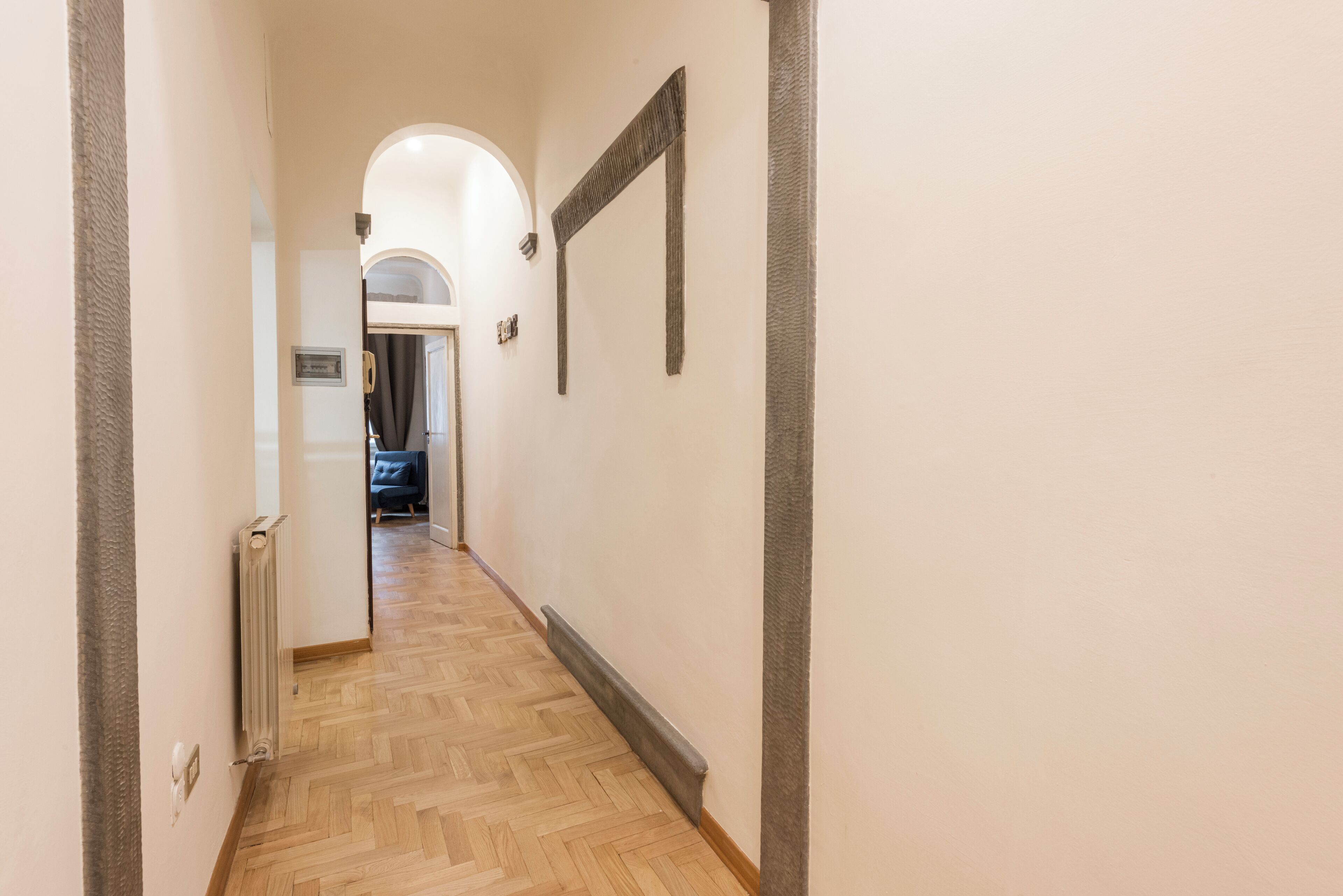 A hallway with a wooden parquet floor, a radiator, and a door at the end leading to a room with a dark-colored sofa.