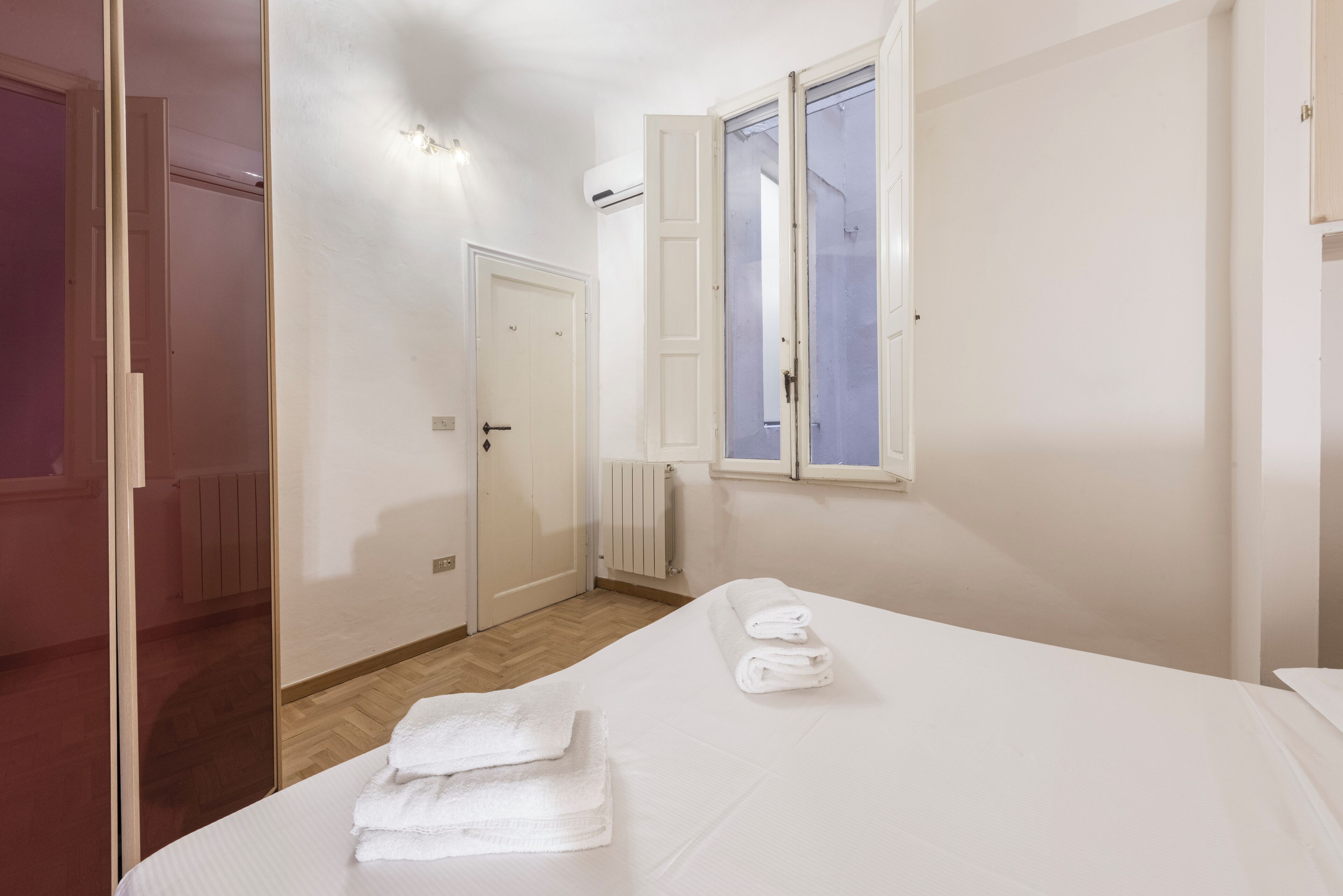 A hotel room with a bed, a window with an open door, a radiator, and a mirror.