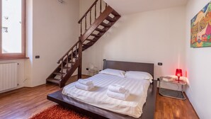 Un dormitorio con una escalera de madera, una cama con ropa blanca, una lámpara roja sobre una mesita de noche y un cuadro en la pared.