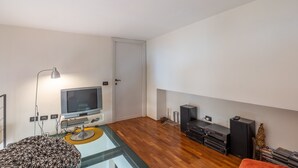 Una sala moderna con televisor de pantalla plana, una lámpara de pie, un reposapiés estampado, una mesa de centro de vidrio, piso de madera, un mueble de entretenimiento negro y un pequeño árbol de Navidad.