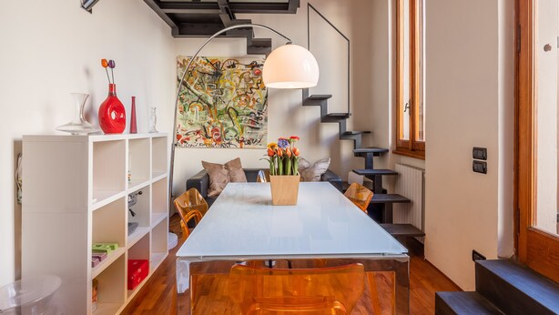 Un comedor moderno con una mesa blanca, sillas de madera, un florero con flores, un cuadro abstracto y colorido, y una escalera.