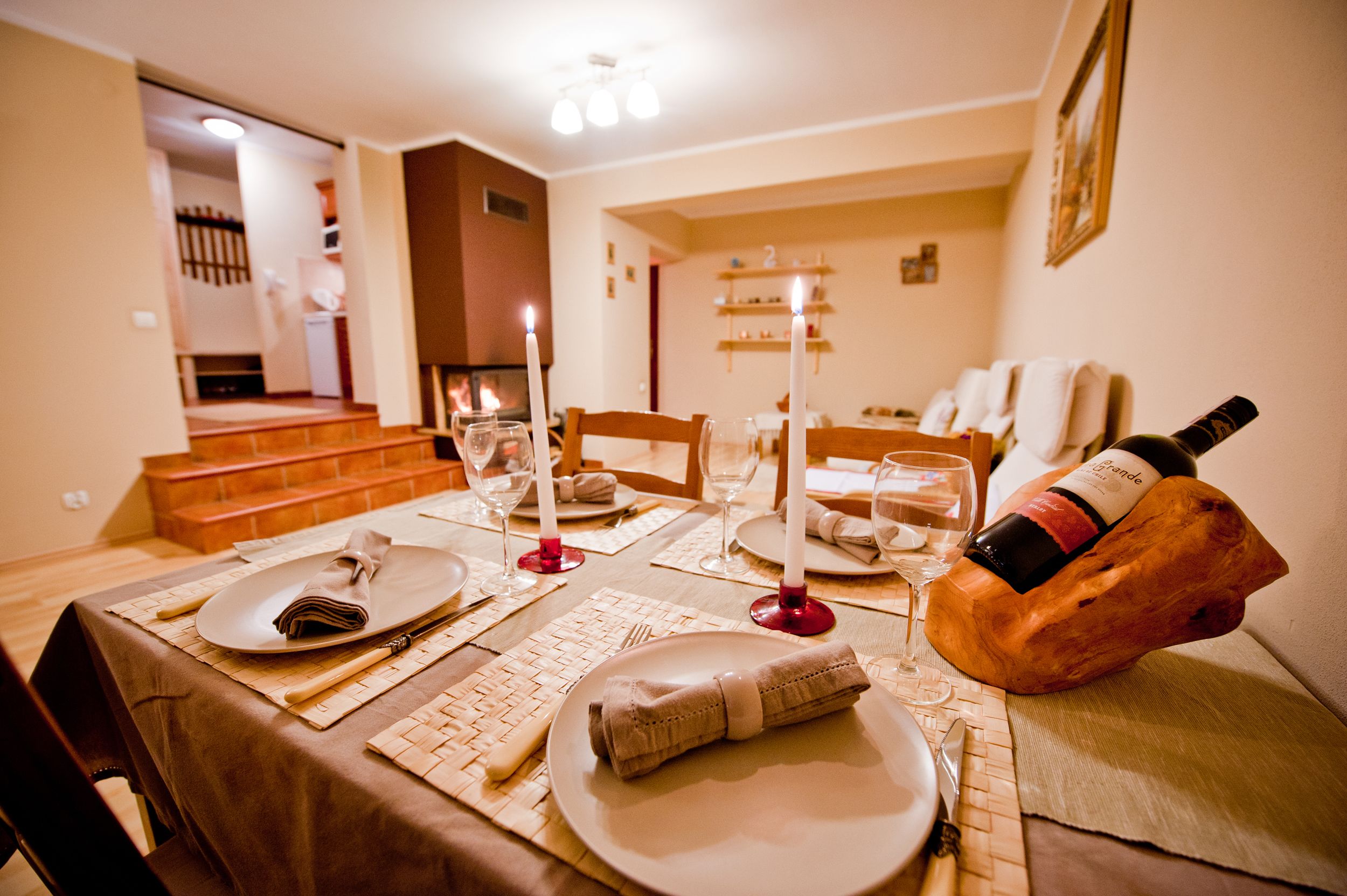 Une table à manger dressée avec des assiettes, des couverts et des verres, et une bouteille de vin posée sur un bloc de bois.