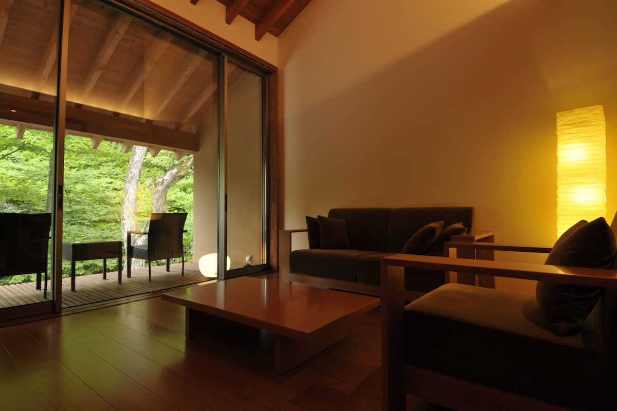 A modern living room with a sofa, coffee table, and chairs, a large window with a view of greenery, and a wall-mounted lamp.