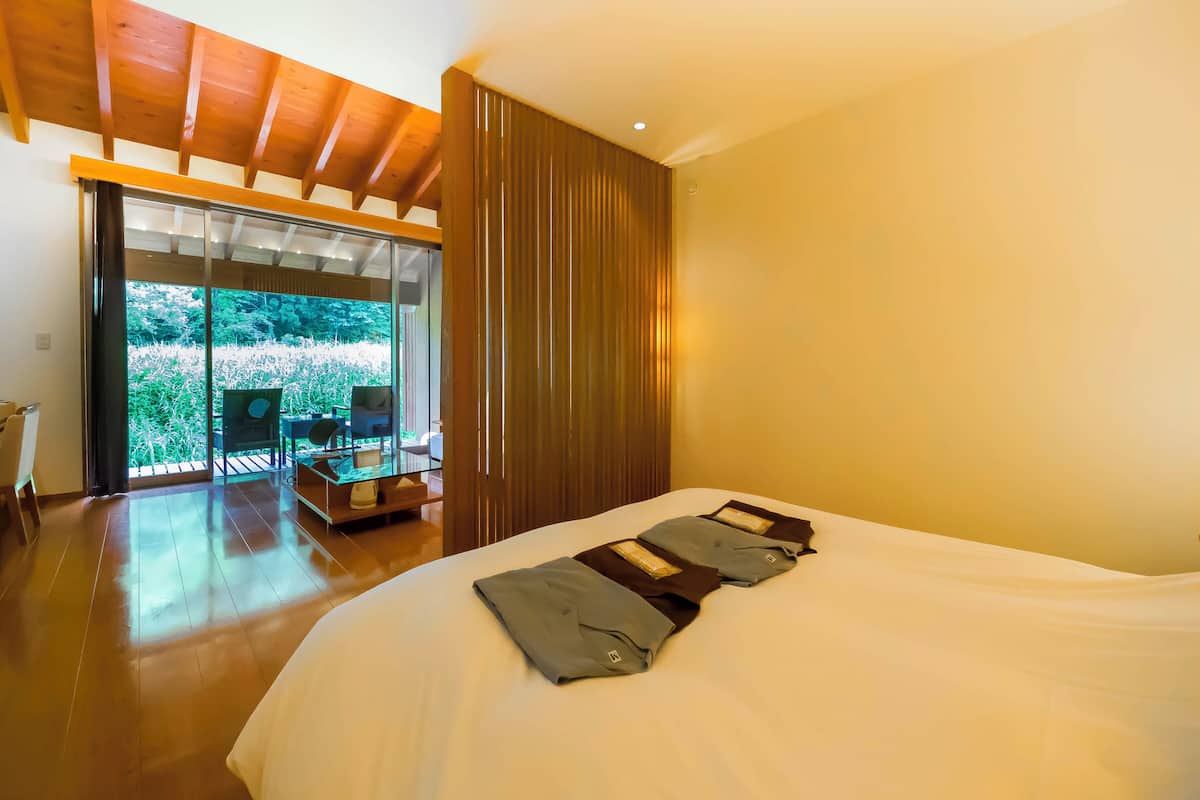 A hotel room with a large bed, wooden flooring, a sliding glass door leading to an outdoor area, and a wooden ceiling.