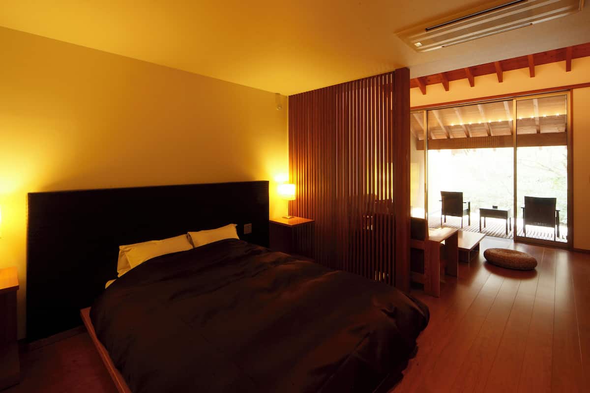 A bedroom with a large bed, a nightstand with a lamp, a wooden partition, and a view of an outdoor deck.
