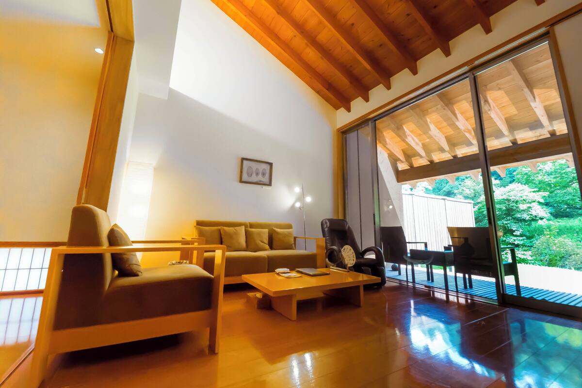 A modern living room with a sofa, coffee table, and chairs, large sliding glass doors leading to a deck with a view of greenery.