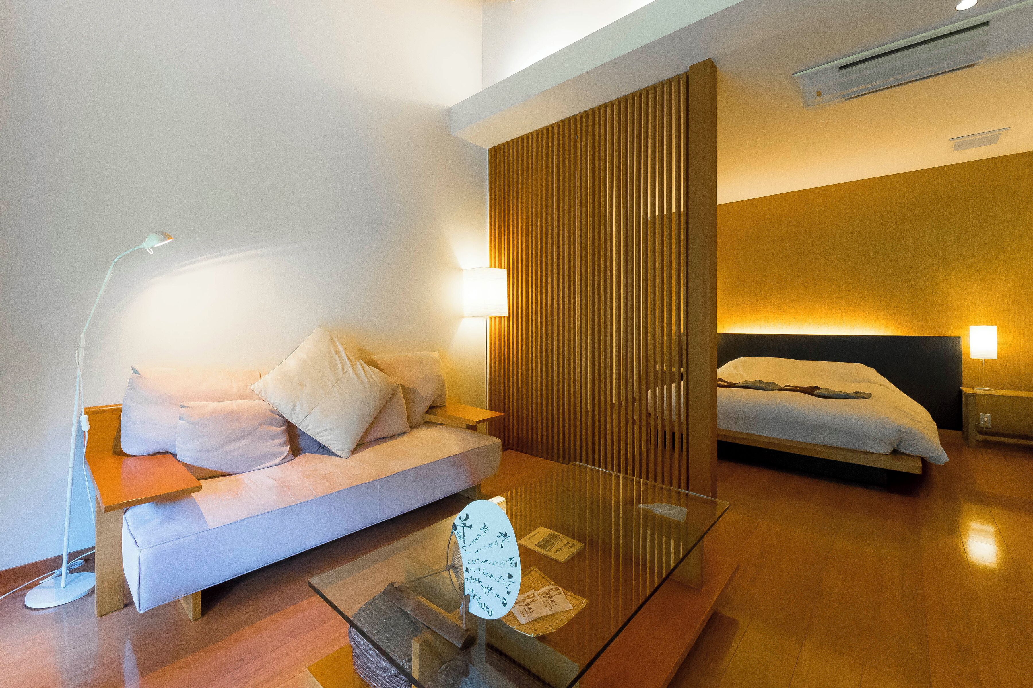 A modern hotel room with a sofa, a glass coffee table, a bed, and a wooden partition.