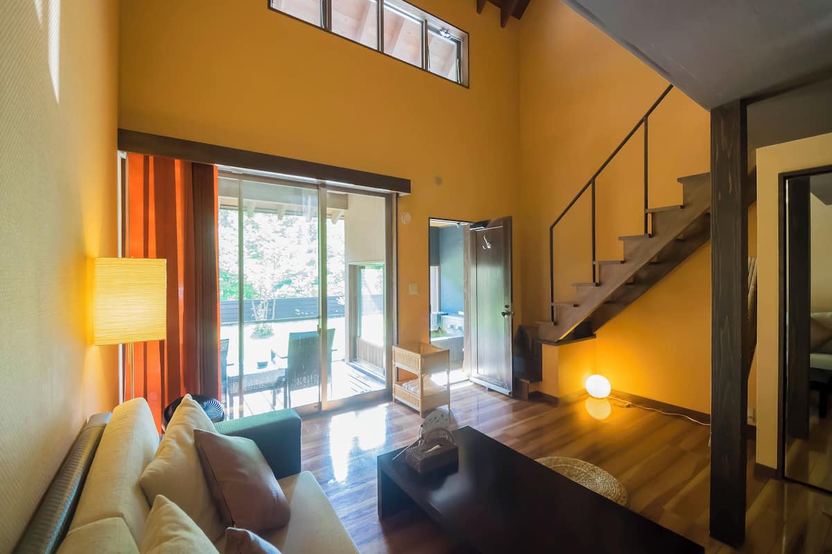 A spacious living area with a sofa, a coffee table, and a lamp. There is a staircase leading to an upper level, and a sliding glass door opens to a balcony.