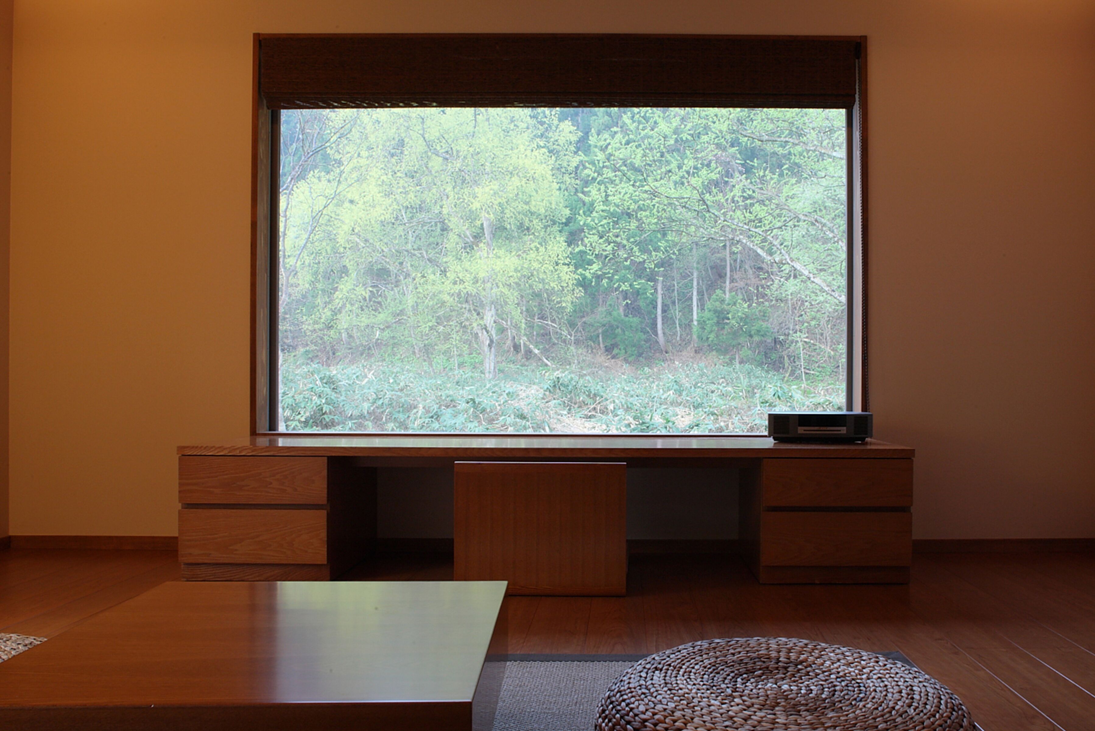 A room with a large window overlooking a forest, a wooden desk, and a woven floor mat.