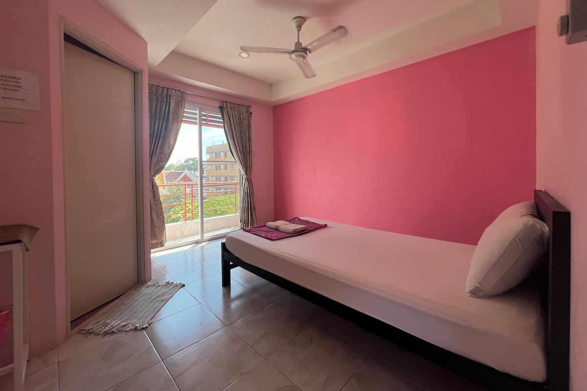 A bedroom with a bed, a ceiling fan, a window with curtains, and a view of buildings outside.