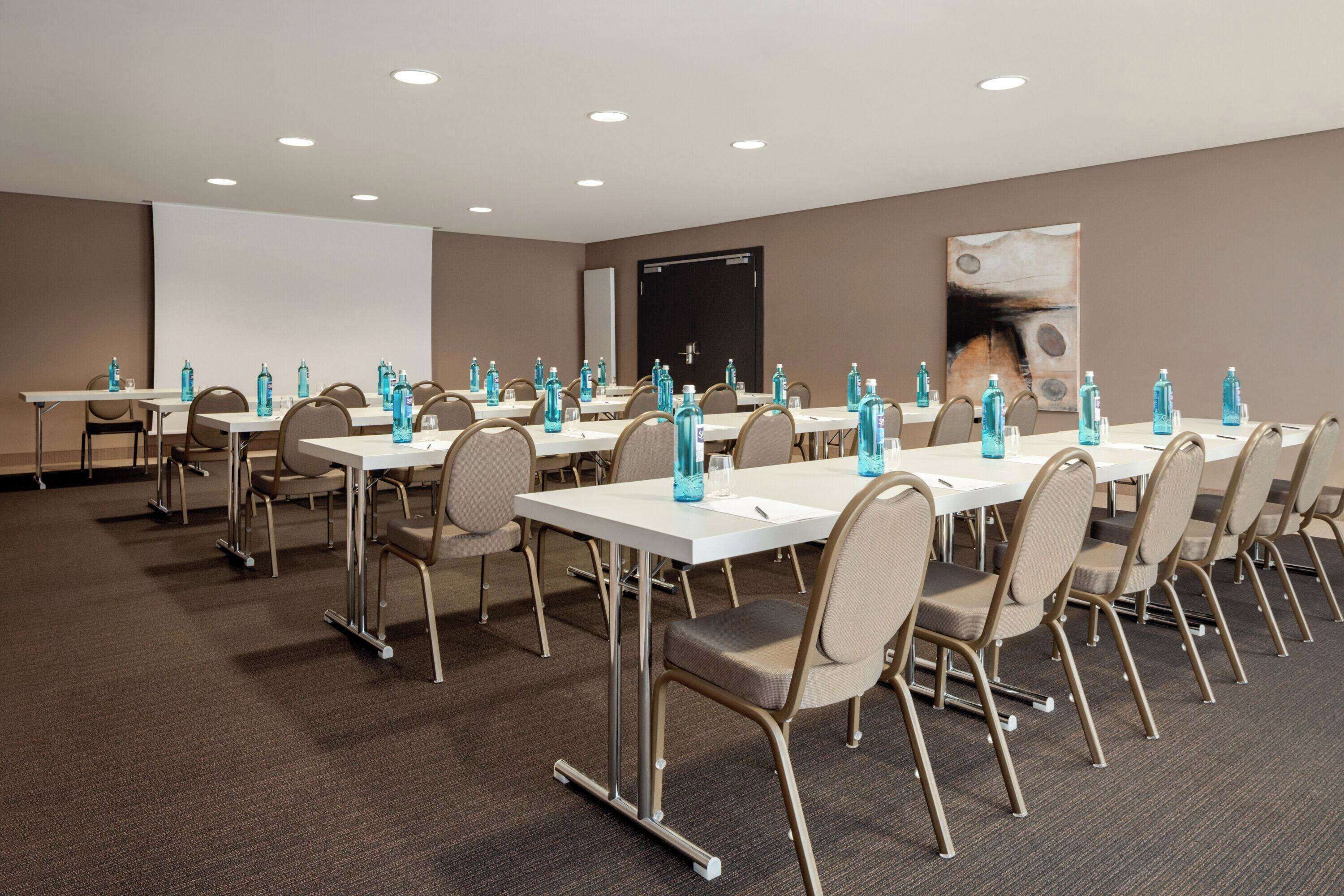 A conference room with multiple tables and chairs, equipped with water bottles and a projector screen.