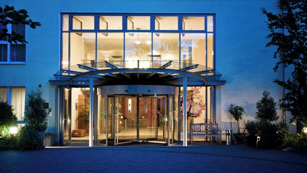 Entrada moderna de hotel com porta giratória de vidro, um banco do lado de fora e interior bem iluminado visível pelas janelas.