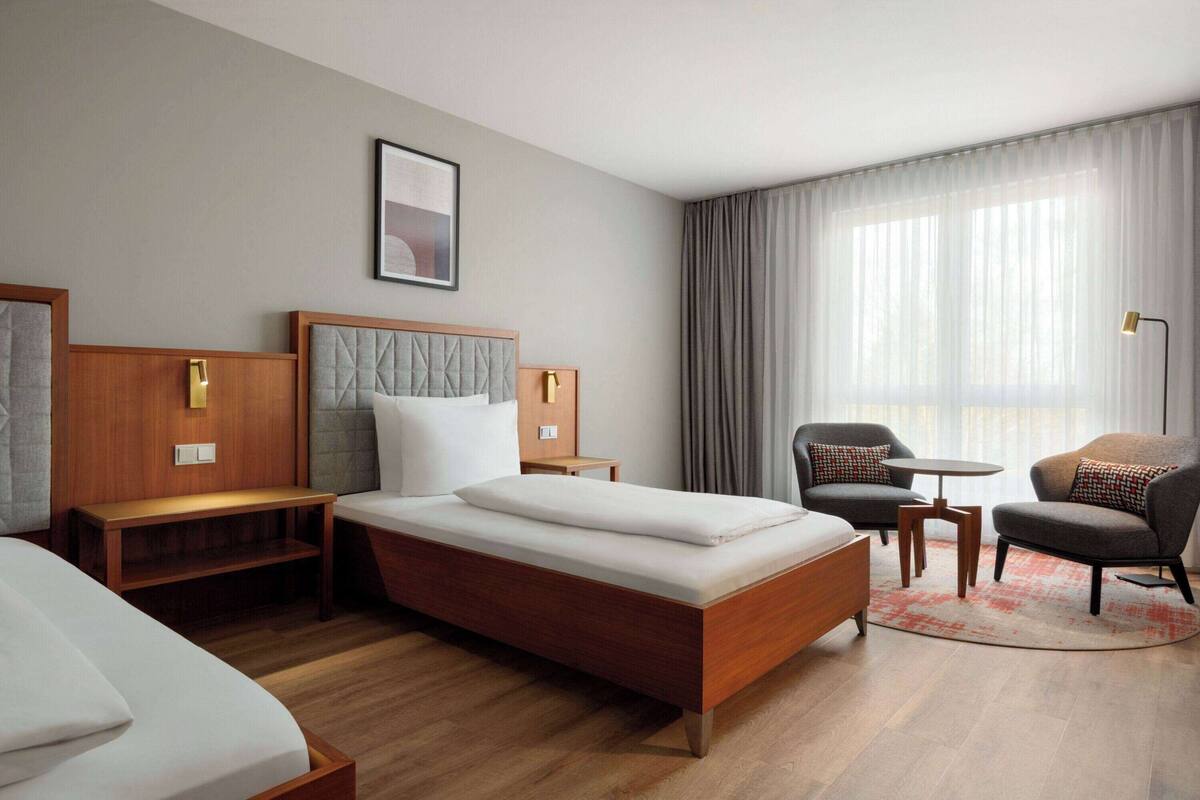 Een hotelkamer met een groot bed, twee nachtkastjes, een stoel, een klein tafeltje en een raam met gordijnen.
