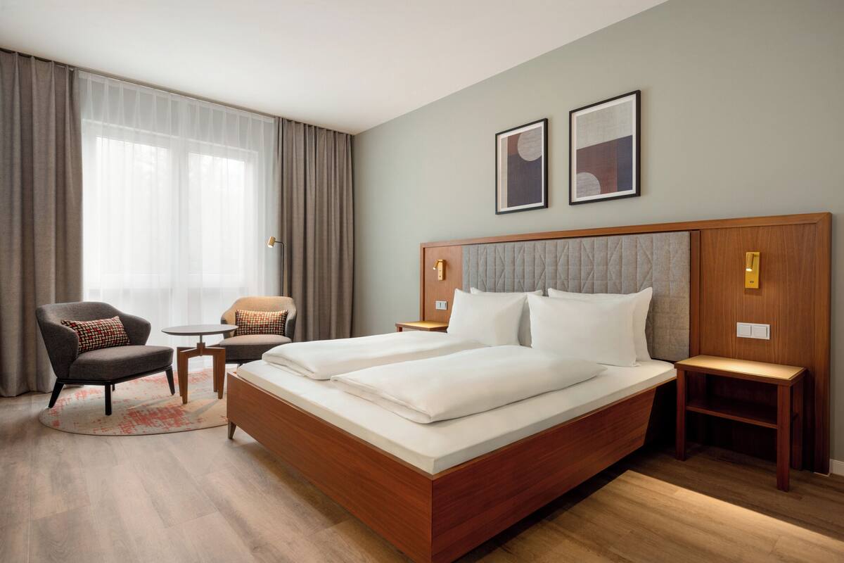 Hotelkamer met een groot bed, twee nachtkastjes, een stoel, een klein tafeltje en een raam met gordijnen.